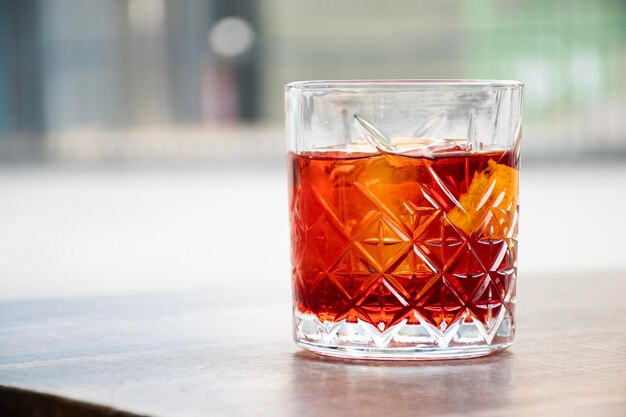 glass of red negroni cocktail with orange slices and clear ice block on wooden table blurred city background