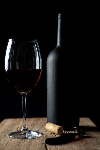 Glass of red wine on the rustic wooden table, with corkscrew beside it and defocused wine bottle