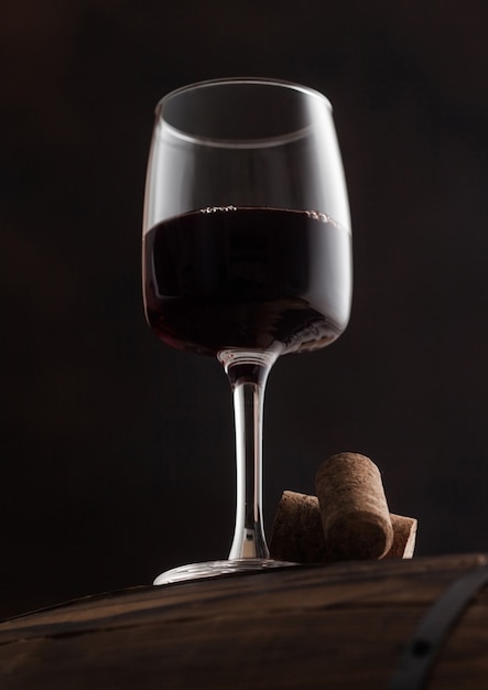 Glass of red wine with corks on top of wooden barrel on black background.