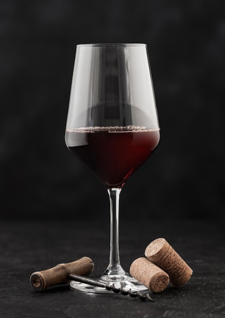 Glass of red wine with corks and vintage corkscrew on dark background