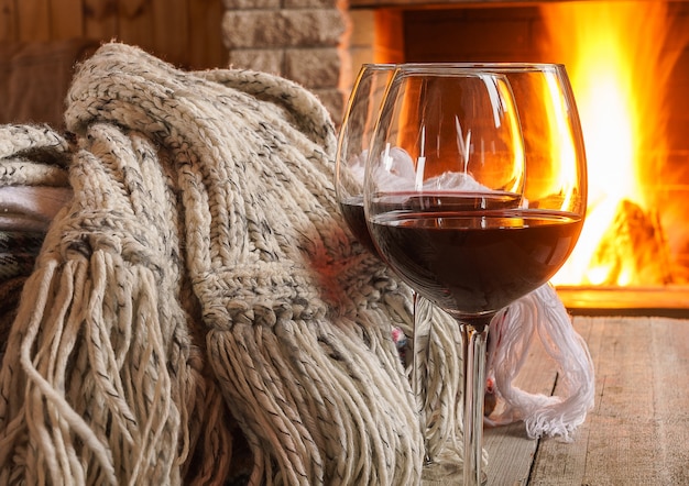 Glass of red wine and wool things near cozy fireplace. 