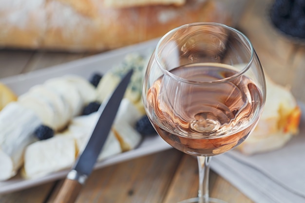 A glass of rose wine served with cheese plate, blackberries and baguette. Assortment of cheese with berries on wooden background.