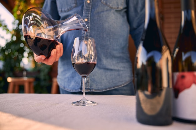 Bicchiere sul tavolo e un uomo in camicia di jeans che ci versa del vino rosso dal decanter