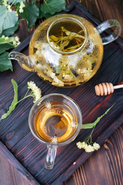 Стеклянный чайник и чашка липового чая на темном деревянном подносе на столе, здоровый травяной чай, крупным планом.