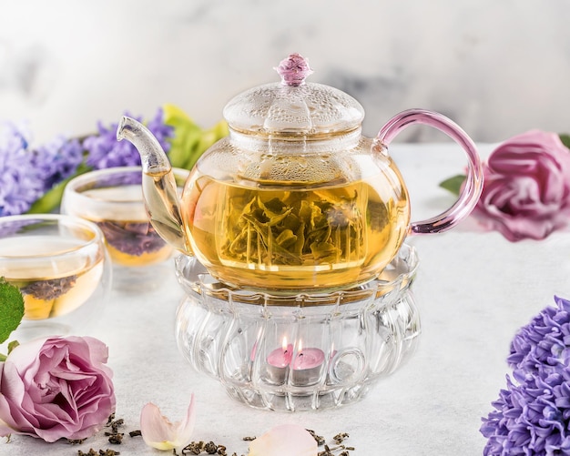 Glass teapot and cups with green tea on light background decorated with flowers