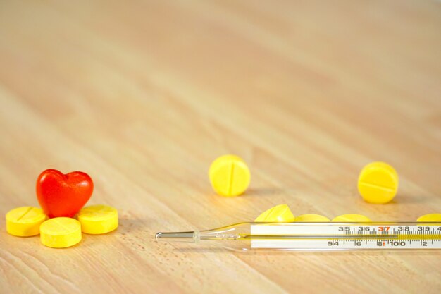 Glass thermometer with yellow medicines and mini red heart on wood table.