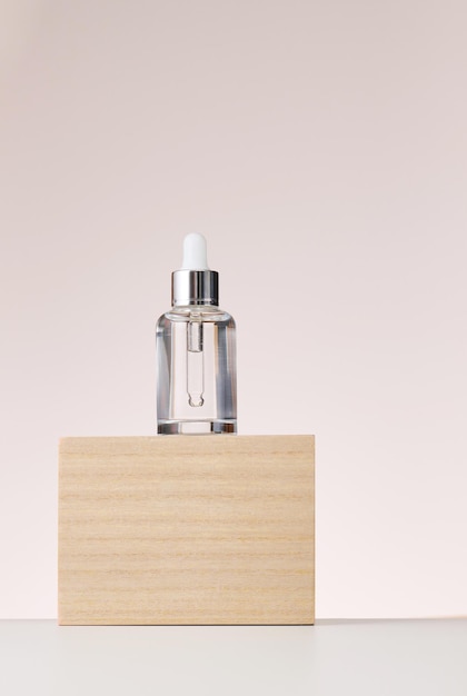 Стеклянная прозрачная белая бутылка с пипеткой стоит на деревянном подиуме Контейнер для косметических кислот и масел на бежевом фоне