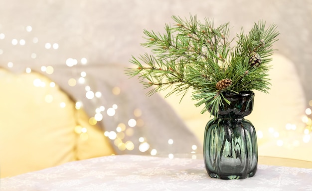 Glass vase with fir branches on table with tablecloth in living room with sofa and Christmas lights