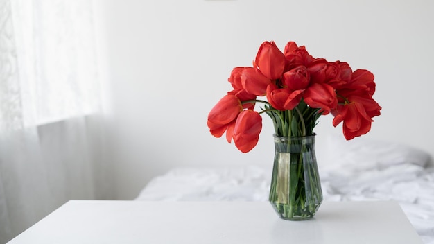 Glass vase with red tulip flowers in a light cozy bedroom interior White wall bed with white linen Banner Home vacation place