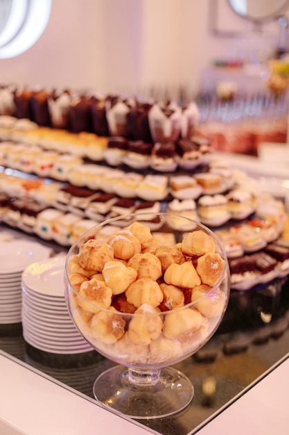 Glass vase with small cakes stands on dessert table, behind it\
are various cakes and desserts. wedding event. service at hotel.\
recreation and travel. sweet buffet.