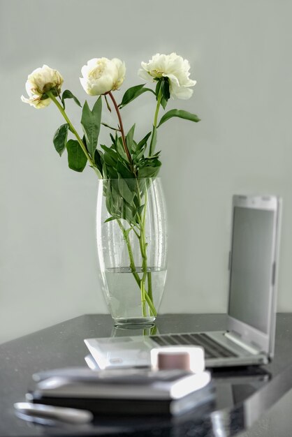 写真 ラップトップを開いて作業場所に立つ白い牡丹のガラス花瓶