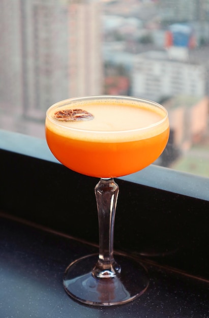 Glass of vibrant orange cocktail at rooftop terrace with blurry urban view in the backdrop