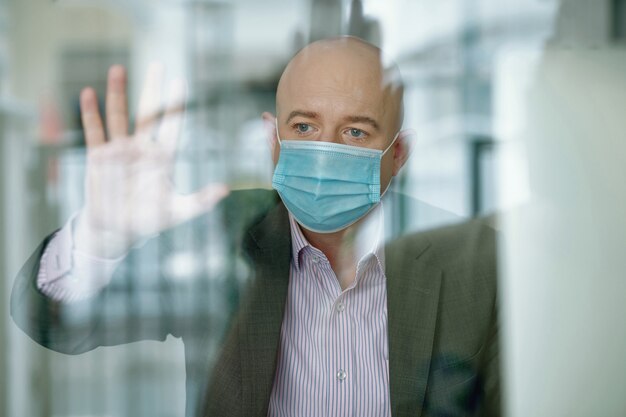 Behind glass view of sad mature businessman in mask touching glass box while being in isolation because of coronavirus pandemic