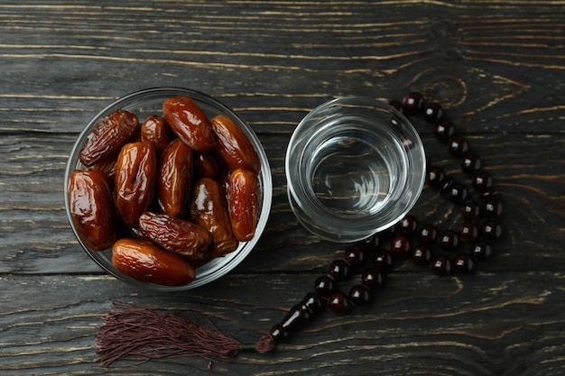 Glass of water, bowl of dates and rosary on wooden surface