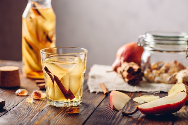 Glass of Water Infused with Sliced Pear, Cinnamon Stick, Ginger Root and Some Sugar. Ingredients on Wooden Table and Bottle of beverage on Backdrop.