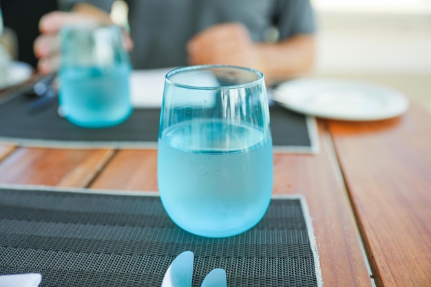 A glass of water on a table with a man sitting at the table in the background.