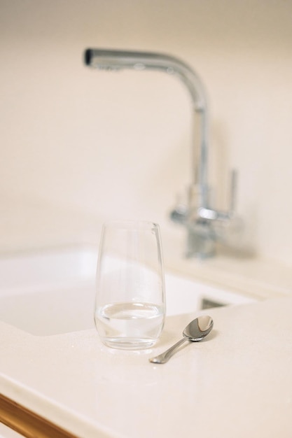 Glass of water and teaspoon on table in kitchen with faucet closeup