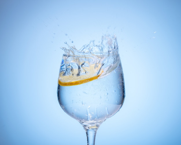 Bicchiere d'acqua con limone caduto e spruzzata su sfondo azzurro sfumato.