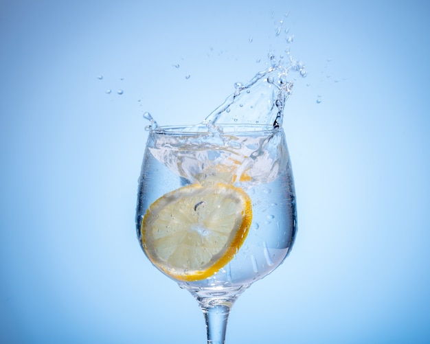 Glass of water with lemon droped in and splash on light blue gradient background.