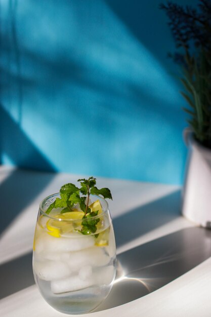 glass of water with mint, lemon and ice on white table, blue background