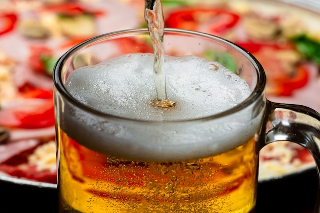 A glass in which beer is poured close-up on a background of pizza.