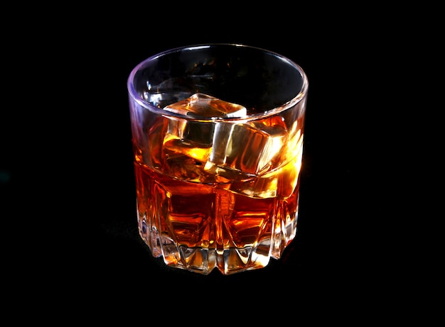 Photo glass of whiskey or bourbon with ice on black stone table.