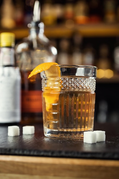 Glass of whiskey cognac or brandy with lemon and ice cubes standing on the bar counter with a bottle on the background