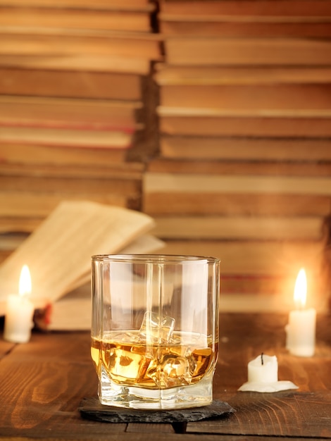 Un bicchiere di whisky sul tavolo con libri e candele accese e raggi di luce. copia spazio, foto verticale.