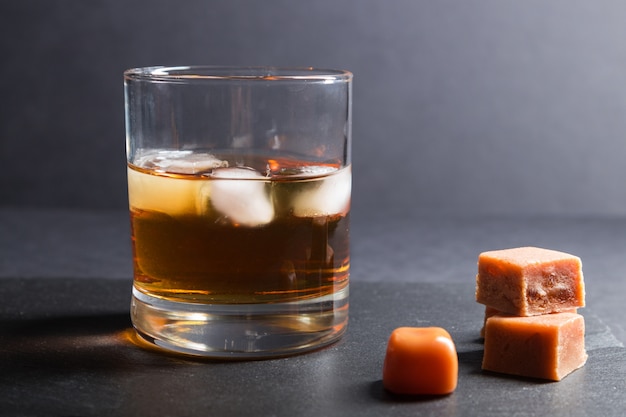 Glass of whiskey with ice and caramel candies on a black stone slate board on black surface. Side view, selective focus.