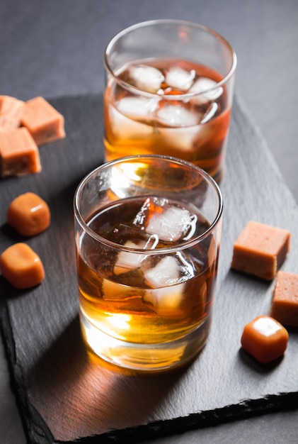 Glass of whiskey with ice and caramel candies on a black stone slate board on black surface. Side view, selective focus.