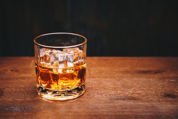 Glass of whiskey with ice cube on wooden table over black background