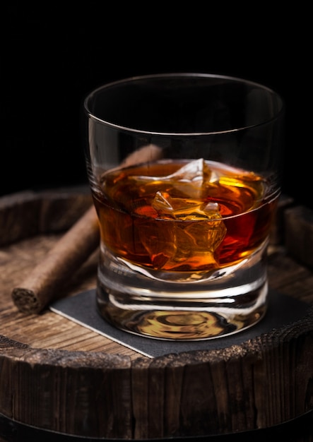 Glass of whiskey with ice cubes and cigar on top of wooden barrel