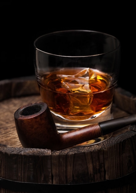 Bicchiere di whisky con cubetti di ghiaccio e tubo di fumo vintage in cima a botte di legno