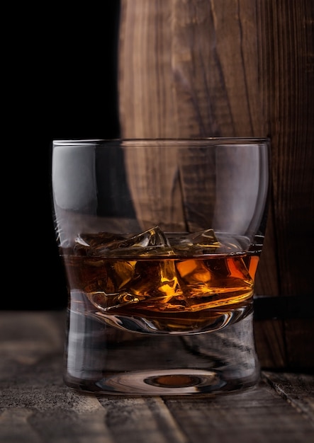 Glass of whiskey with ice cubes next to wooden barrel   