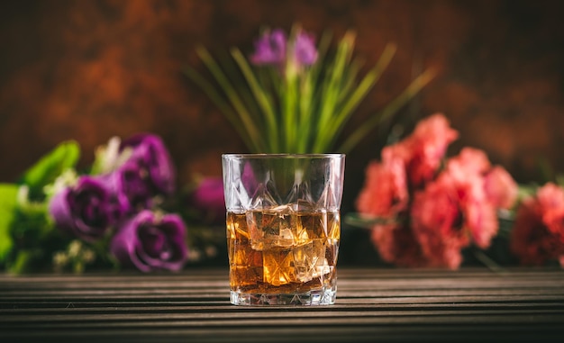 Glass of whiskey with ice on a wooden table with some color flowers