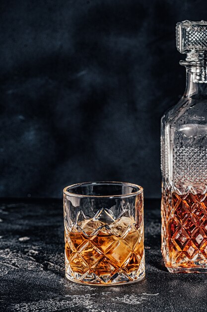 Glass of the whiskey with a square decanter on a black stone background.