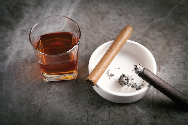 Стакан виски, пепельница и сигары на столе.