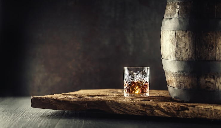 Photo glass of whisky cognac or bourbon in ornamental glass next to a vinatge wooden barrel on a rustic wood and dark background.