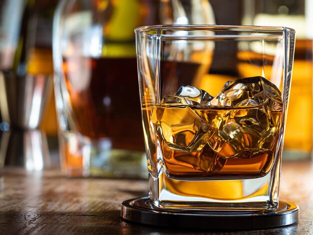 Glass of whisky on the rocks on a wooden bar top with bottles in the background