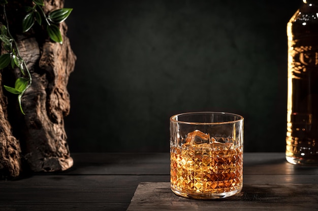 Photo glass of whisky with ice and bottle on wooden table on dark background with copy space