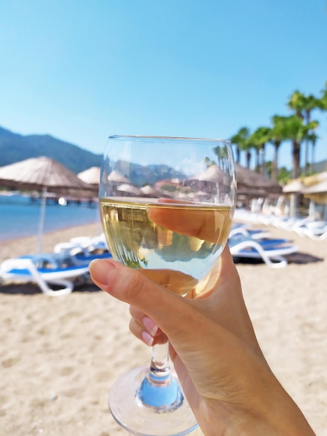 a glass of white wine in hand on the background of a sunny beach with sun loungers and umbrellas