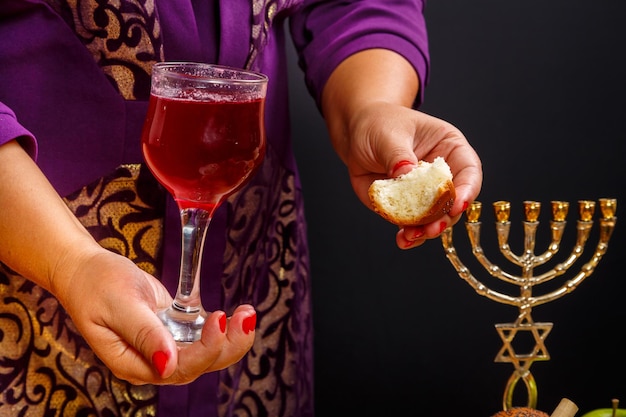 A glass of wine and a piece of challah in women's hands on the day of Rosh Hashanah in women's hands over a table with a menorah