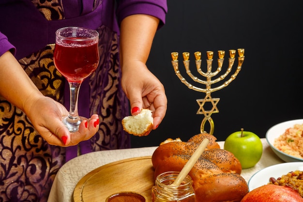 A glass of wine and a piece of challah in women's hands kiddush on the day of Rosh Hashanah in women's hands over a festive table with a menorah