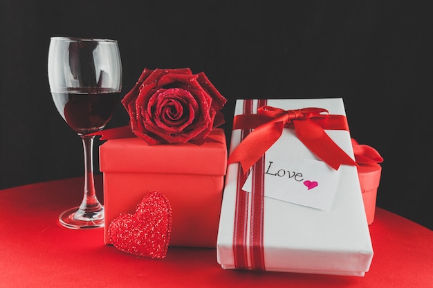 Foto bicchiere di vino con regali e una rosa su un tavolo rosso