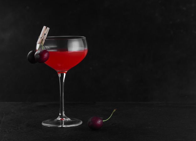 A glass with an alcoholic cocktail of red color decorated with cherries on a clothespin. Low key