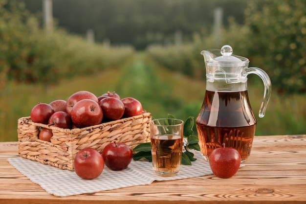 A glass with apple juice and basket with apples on wooden table with natural orchard background Vegetarian fruit composition