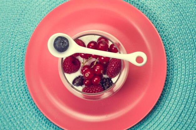 A glass with berries and yogurt and a spoon with a blueberry on a pink plate in a top view