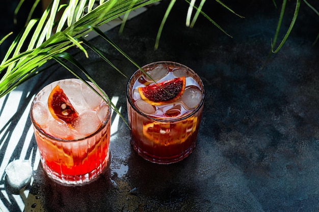 Glass with cold mineral water with blood oranges and ice cubes on concrete background Tropical palm leaf shadow Sunlight and shadow background