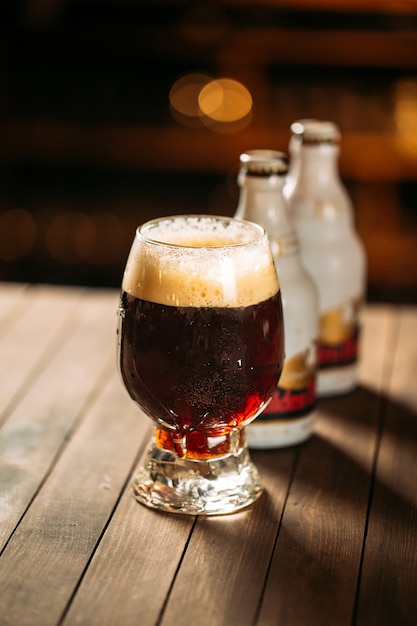 Glass with a dark foamed beer on the wooden table with two beer bottles