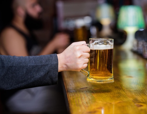 거품과 신선한 라거 생맥주와 유리입니다. 남성의 손은 바에서 차갑고 맛있는 맥주로 가득 찬 머그를 들고 있습니다. 금요일 여가 전통. 맥주 펍 개념입니다. 바 카운터 defocused 배경에 맥주 잔입니다.